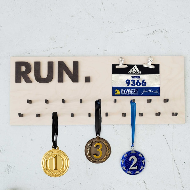 May include: A wooden medal display with the word "RUN." printed in black. The display has 24 hooks for hanging medals. Three medals are hanging on the hooks. The medals are gold, bronze, and blue. A blue and black marathon race bib is attached to the display with two silver clips. The bib reads "adidas SIMON 9366 124th Boston Marathon Virtual Experience John Hancock."