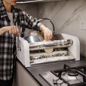 Könnte beinhalten: Eine Person verwendet eine weiße und silberne manuelle Nudelmaschine, um Teig zu flach zu rollen. Die Maschine hat einen Kurbelgriff und ein Tablett aus Metall mit einer mit Mehl bedeckten Oberfläche.