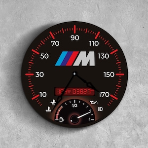 May include: A black round wall clock with a speedometer design. The clock features red markings for speed, a digital display, and the letters "M" and "BMW" in the center. The clock's hands indicate the time.