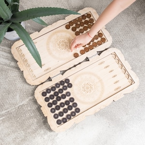 Könnte beinhalten: Ein Backgammon-Set aus Holz mit aufwendigen Schnitzereien. Das helle Spielbrett enthält dunkelbraune und hellbraune Spielsteine. Eine Hand platziert einen Spielstein. Das Set steht auf einer grauen Oberfläche.