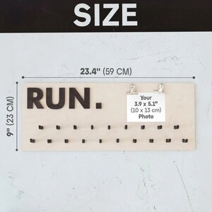 May include: A wooden medal display board with the word "RUN." in large black letters. The board has 40 small hooks for hanging medals. The board measures 23.4 inches wide and 9 inches tall. A small white card with the text "Your 3.9 x 5.1" (10 x 13 cm) Photo" is attached to the board with two silver clips.
