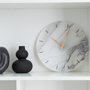 May include: A round wall clock with a white marble face and gold-colored hands. The clock has black dashes marking the hours. Two black decorative vases are on the left side of the clock. The vases are black and have a glossy finish.