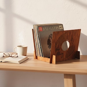 Puede incluir: Un soporte de discos de madera con un diseño de vinilo cortado con láser, que contiene varios discos de vinilo. El soporte es de madera marrón claro y está sobre una mesa de madera. Una taza de café, un libro abierto y gafas también están sobre la mesa.