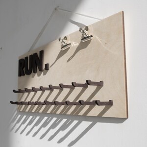 May include: A wooden wall-mounted rack with the word "RUN." in black letters. The rack has 20 hooks for hanging items, such as running shoes or medals.