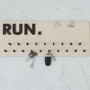 May include: A wooden key holder with the word "RUN." printed in black letters. The holder has 28 small hooks for hanging keys. There are two silver clips at the top of the holder.