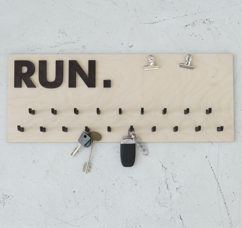 May include: A wooden key holder with the word "RUN." printed in black letters. The holder has 28 small hooks for hanging keys. There are two silver clips at the top of the holder.