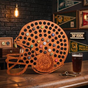 May include: A wooden football helmet-shaped bottle cap holder with numerous holes and a few bottle caps inserted. The holder has the text "WILLIAM'S Beer EST 2001" engraved on it. A glass of beer and a bottle opener are on the wooden surface.