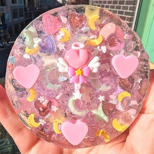 May include: A clear pink resin coaster with a pink heart, a wand with a pink heart and wings, and a yellow crescent moon. The coaster is filled with glitter and other small pink and yellow decorations.