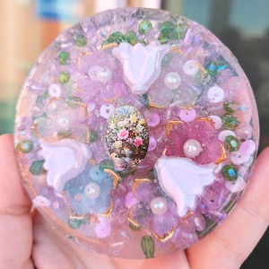 May include: A round, clear resin coaster with a floral design. The coaster features pink, white, and green flowers, as well as gold accents. A small, framed image of a floral bouquet is in the center of the coaster.