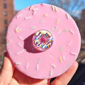 Puede incluir: Un cenicero de vidrio en forma de rosquilla rosa con una rosquilla de vidrio más pequeña en el centro. La rosquilla más pequeña tiene glaseado blanco y chispas de colores. La rosquilla más grande tiene chispas blancas.