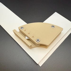 K&ouml;nnte beinhalten: Ein beigefarbener Papierschneider mit Klinge, der auf wei&szlig;em Papier liegt. Der Schneider hat ein gebogenes Design mit drei kleinen Dreiecken und drei silbernen Schrauben. Das Papier ist in V-Form vor einem schwarzen Hintergrund angeordnet.