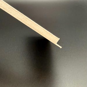 K&ouml;nnte beinhalten: Ein Abschnitt einer hellfarbigen Holzleiste, diagonal vor einem dunklen Hintergrund. Die Leiste hat ein rechteckiges Profil mit einer kleinen Lippe an einer Kante, wahrscheinlich zur Installation. Die Holzmaserung ist sichtbar.