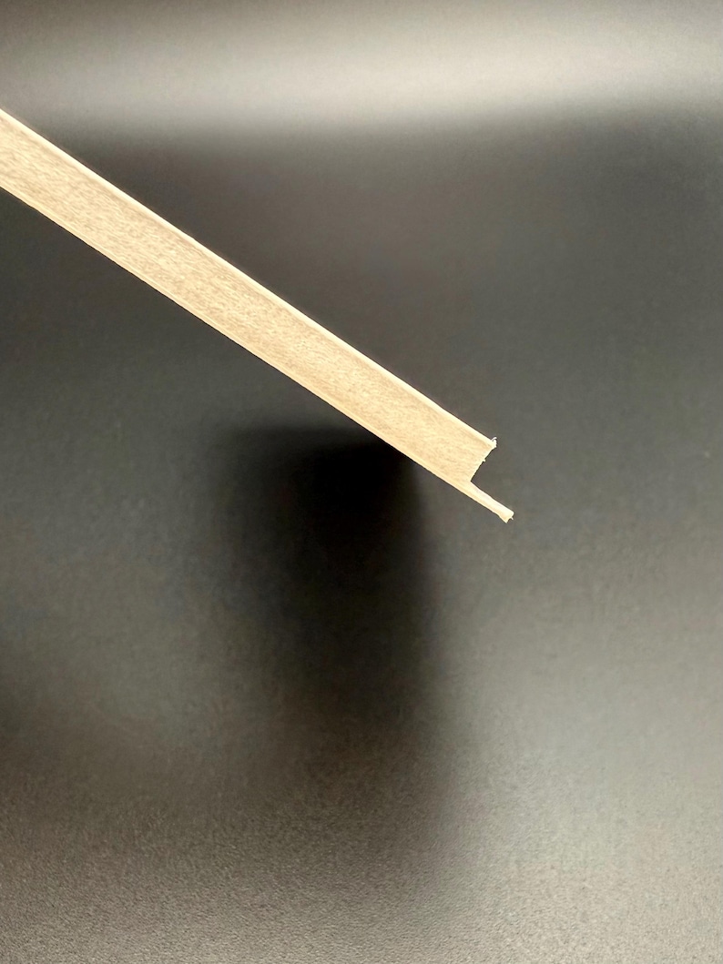 K&ouml;nnte beinhalten: Ein Abschnitt einer hellfarbigen Holzleiste, diagonal vor einem dunklen Hintergrund. Die Leiste hat ein rechteckiges Profil mit einer kleinen Lippe an einer Kante, wahrscheinlich zur Installation. Die Holzmaserung ist sichtbar.