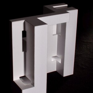K&ouml;nnte beinhalten: Eine wei&szlig;e, geometrische Papierskulptur mit minimalistischem Design. Die Skulptur weist rechteckige und quadratische Ausschnitte auf, die einen dreidimensionalen Effekt erzeugen. Das Kunstwerk ist vor einem schwarzen Hintergrund platziert.