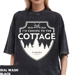 May include: Black mineral wash t-shirt with a white graphic. The graphic reads "I'M COMING TO THE COTTAGE" with a shield design, trees, and the word "CANADA". The shirt has short sleeves.