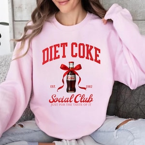 Puede incluir: Una sudadera rosa claro con las palabras "DIET COKE" en rojo en forma de arco. Debajo hay un gráfico de una botella de Coca-Cola Light con un lazo rojo, el texto "EST. 1982" y "Social Club" con el lema "JUST FOR THE TASTE OF IT".