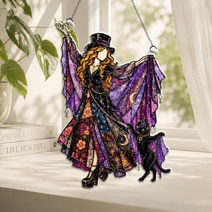 May include: A stained glass-style hanging ornament featuring a figure in a top hat and flowing purple cape with celestial patterns, standing next to a black cat with wings. The figure wears a long dress with floral and star motifs and platform shoes.