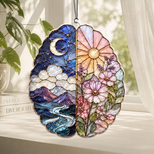 Peut inclure: Un attrape-soleil en vitrail en forme de cerveau avec un design double. Le côté gauche représente une scène nocturne avec un croissant de lune, des nuages, des montagnes et une rivière dans des tons bleus et violets. Le côté droit montre un soleil.