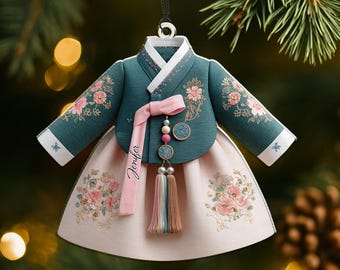 Könnte beinhalten: Ein teal- und rosa Hanbok-Ornament, ein traditionelles koreanisches Kleid, hängt an einem Faden. Das Ornament hat Blumenstickereien, ein rosa Band mit dem Namen "Jenifer" und eine Quaste. Der Hintergrund ist verschwommen mit Tannenzweigen.