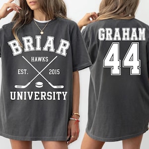 May include: Dark gray t-shirt with the text "BRIAR HAWKS UNIVERSITY" and crossed hockey sticks. The back of the shirt displays the name "GRAHAM" and the number "44" in white.