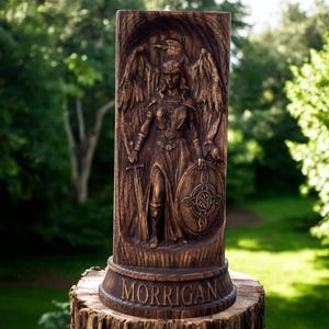 May include: A carved wooden statue of Morrigan, a figure with wings, a sword, and a shield. The statue is brown and stands on a wooden base with the name "MORRIGAN" carved into it. The background is a blurred green forest.