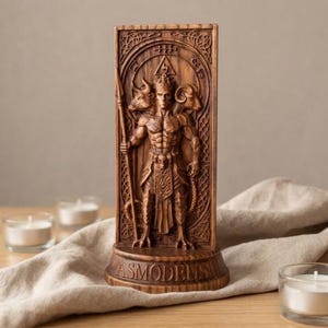 May include: A rectangular brown wooden plaque featuring a relief carving of a central figure with three heads, including a bull and a ram, holding a spear. The base is inscribed with the name ASMODEUS in capital letters within a decorative border.