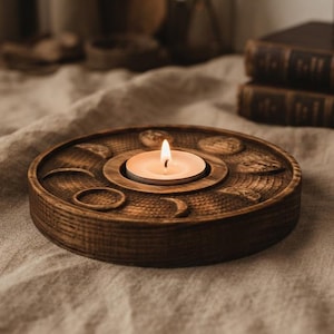 May include: A round, dark-stained wooden candle holder featuring carved moon phase symbols arranged in a circular pattern around a central recessed area. A single white tea light candle sits in the center with a small, glowing yellow flame.