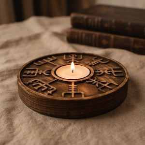 May include: A circular dark brown wooden candle holder featuring carved runic symbols arranged in a radial pattern around a central circular opening. A small white tea light candle is placed inside the opening with a lit flame visible.