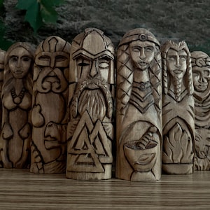 May include: A set of nine wooden carvings depicting Norse deities and symbols. The carvings are detailed and intricate, with each figure featuring unique facial expressions and clothing. The carvings are arranged in a row on a wooden surface.