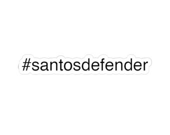 Peut inclure: Autocollant blanc avec le texte "#santosdefender" en noir, police sans-serif. L'autocollant a une bordure blanche et est conçu pour être appliqué sur diverses surfaces.
