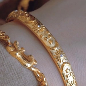 May include: A gold bangle bracelet featuring an embossed pattern of suns, moons, and stars with small inset clear stones, paired with a gold chain bracelet featuring a sculpted animal figure with a textured surface and a small clear stone.