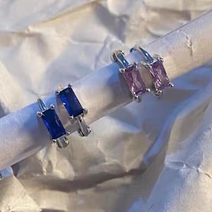 May include: Four silver hoop earrings featuring rectangular baguette-cut gemstones. Two earrings are set with deep blue stones, while the other two earrings are set with light purple stones. Each earring has a hinged closure mechanism.