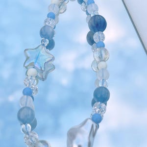 May include: A blue and white beaded necklace with a star charm and a pearlescent teardrop pendant. The necklace is reflected in a mirror.
