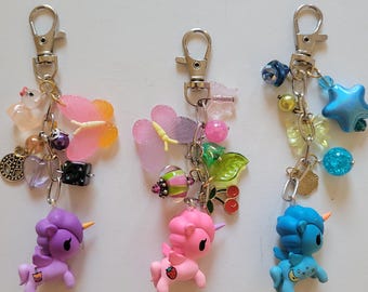 May include: Three metal keychains featuring colorful unicorn charms in purple, pink, and blue. Each keychain includes a silver lobster clasp and a variety of dangling charms, including butterflies, beads, stars, cherries, and small decorative icons.