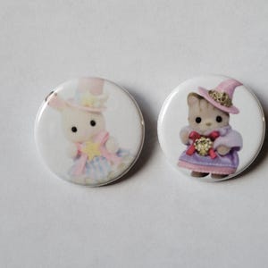 May include: Two round white buttons featuring illustrations of adorable animal characters. One button shows a bunny in a hat and dress, the other a cat in a witch hat and dress. The buttons have a glossy finish and are set against a plain white background.