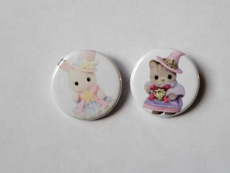 May include: Two round white buttons featuring illustrations of adorable animal characters. One button shows a bunny in a hat and dress, the other a cat in a witch hat and dress. The buttons have a glossy finish and are set against a plain white background.