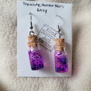 May include: A pair of earrings featuring small glass bottles filled with purple glitter and liquid, each sealed with a cork. The earrings hang from silver-colored hooks. The background includes a white card with the text "Treasure Hunter Nori Etsy".