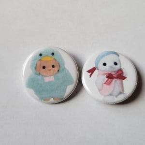 May include: Two round buttons with illustrations. One button features a baby in a blue duck costume. The other button shows a character in a white outfit with a pink scarf and bow. Both buttons have a white background.