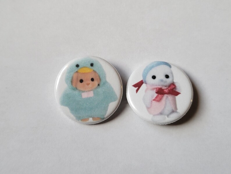 May include: Two round buttons with illustrations. One button features a baby in a blue duck costume. The other button shows a character in a white outfit with a pink scarf and bow. Both buttons have a white background.
