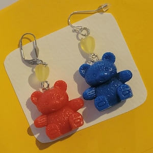 May include: A pair of dangle earrings featuring a blue and a red teddy bear charm. Each charm has a yellow heart bead above it.