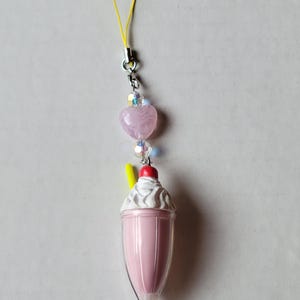 May include: A pink milkshake charm with a white whipped cream topping and a red cherry. The charm has a yellow straw and is attached to a silver chain with a pink heart-shaped bead and iridescent beads. A yellow loop is at the top.