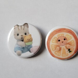 May include: Two round, white buttons. One button features a gray and white cat holding a small yellow cat. The other button has a baby inside a slice of orange. The buttons have a glossy finish.