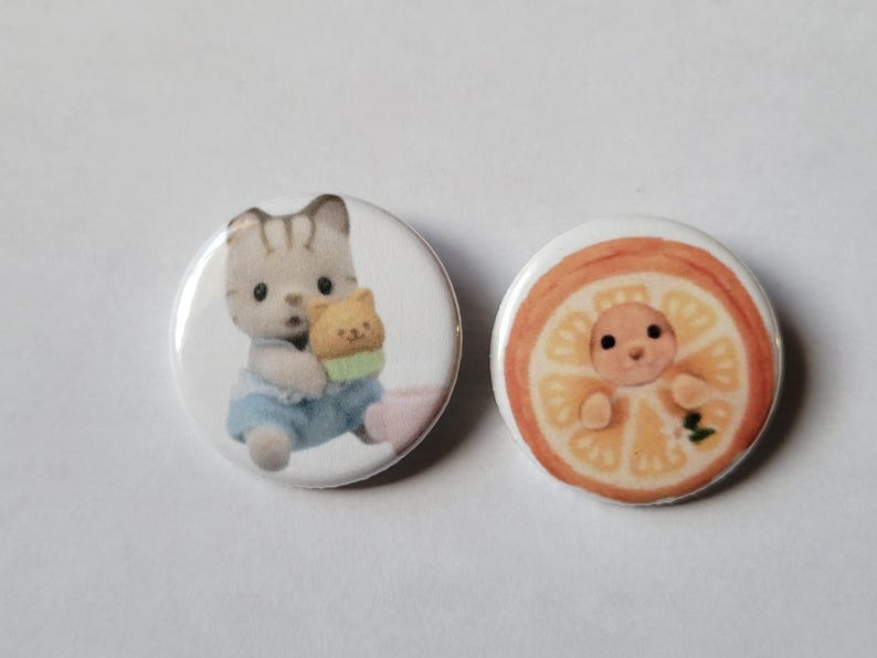May include: Two round, white buttons. One button features a gray and white cat holding a small yellow cat. The other button has a baby inside a slice of orange. The buttons have a glossy finish.