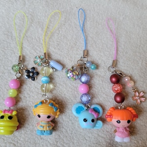 May include: Four colorful charm keychains with beaded accents. Each keychain features a different character: a yellow hippo, a doll, a blue mouse, and a doll with orange hair. The keychains have yellow, blue, and pink cords.