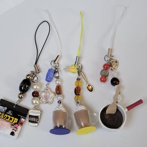 May include: A collection of miniature food-themed charms. Charms include a cocoa packet, coffee cup, whipped coffee mugs, a spoon, and a pan of chocolate. Each charm is attached to a lanyard with beads and other decorative elements.