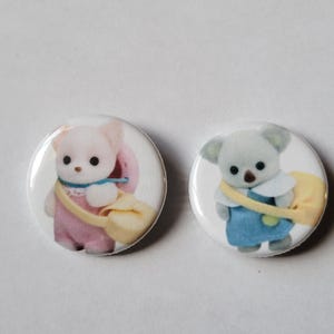 May include: Two round, white buttons featuring cartoon animal characters. One button shows a pink bear with a yellow bag, and the other shows a gray koala with a yellow bag. Both characters are wearing outfits and have black eyes.