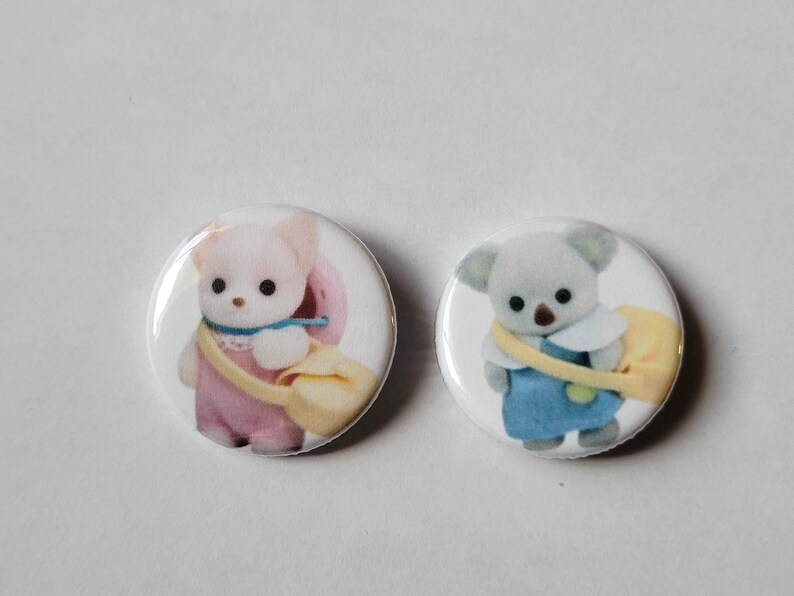 May include: Two round, white buttons featuring cartoon animal characters. One button shows a pink bear with a yellow bag, and the other shows a gray koala with a yellow bag. Both characters are wearing outfits and have black eyes.