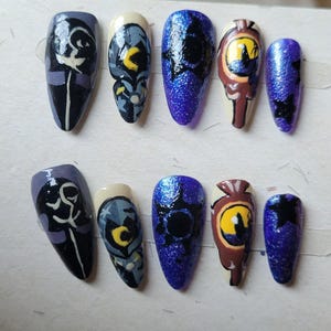 May include: A set of ten artificial nails with various designs. The nails feature a mix of colors, including purple, blue, white, and brown. Some nails have abstract art, while others have a glittery finish or a detailed eye design.