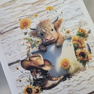 May include: A rustic print featuring a Highland cow peeking over a metal bucket, surrounded by sunflowers. A cowboy hat and boots rest below. The background has a weathered wood texture, creating a charming country aesthetic.