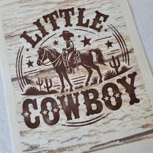 May include: A rectangular card with a brown illustration of a cowboy riding a horse, surrounded by stars and cacti. The words "LITTLE COWBOY" are printed in a bold, brown font, set against a light wood-grain background.